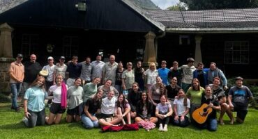 Image of group of Afrikaans male and female high school learners in front of a building sitting and standing on green grass while smiling at the camera.