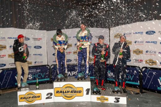 Image of first to third place winners of 5th leg of SA Rally-Raid Championship - 6 men standing on podium holding trophies and spraying champagne.