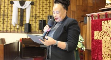 Ms Zulu from the National Department of Transport dressed in black holding a microphone and tablet while delivering a speech in a church.