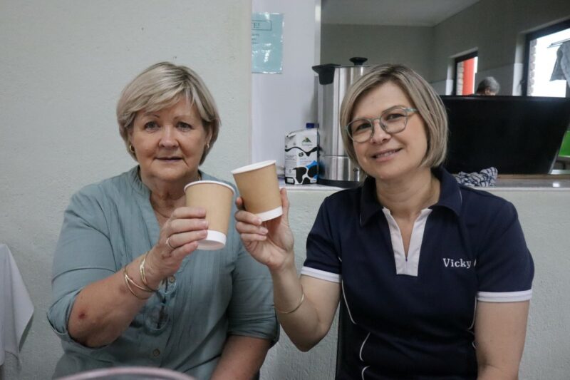 Two woman are sitting behind a table. They are holding brown paper cups in their hands. The older woman is wearing green. The younger woman is wearing navy, with her name embroided in white. There is an urn behind them.