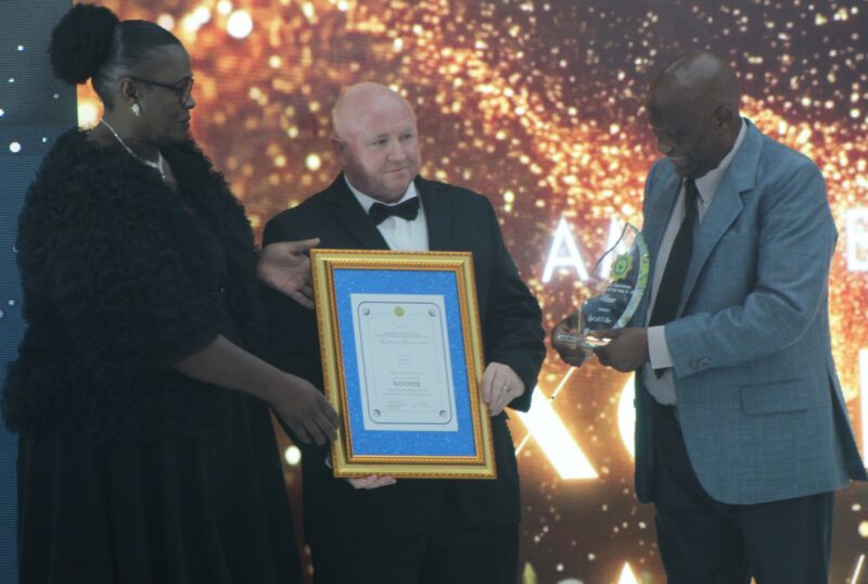 Three people are pictured celebrating an award during an award ceremony. A white, bold man is seen smiling at another woman while both of them are holding a framed certificate, with a second man also in the picture.