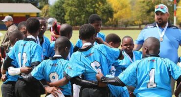 A man is coaching a young rugby team. Uniform is blue and the coach is wearing a blue and white cap.
