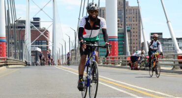 A man joyfully rides his bicycle on a suspension bridge during a cycling event in Johannesburg. He wears a cycling helmet and cycling gear. Other cyclists are pictured in the background.