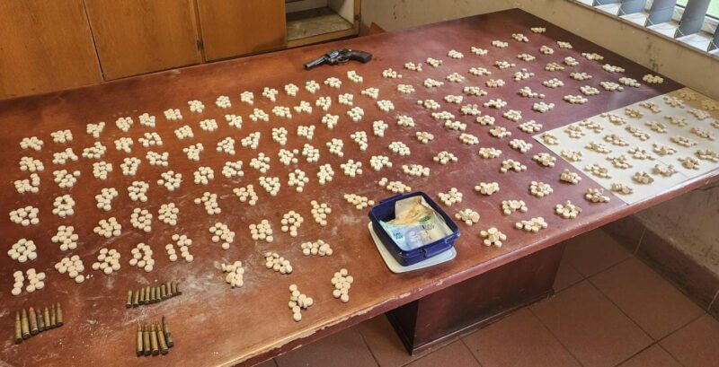 A revolver, several stacks of mandrax drugs, scattered bullet casings, and stacks of assorted money notes are displayed on a wooden table after being seized by the Madadeni Police during a stop-a-search-operation.