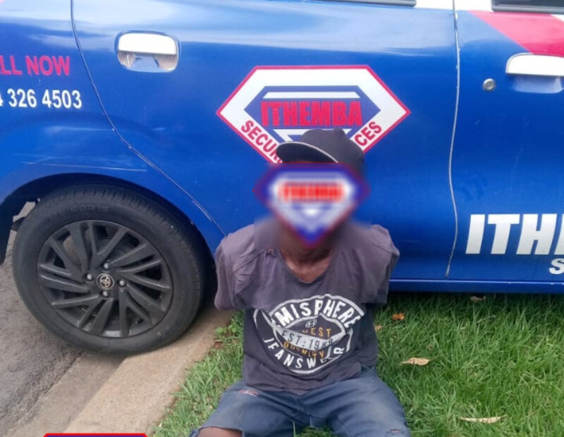 A man sitting on grass, hands behind back, in front of a blue security vehicle with "Ithemba Security Services" logo. The image is of a suspect that has been apprehended by the security team.
