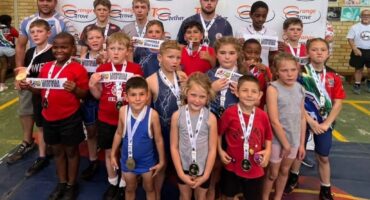 Group of young wrestlers dressed in wrestling attire with medals around their necks and the coaches behind them in front of a Orange Grove Dairy banner.