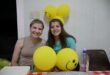 Two girls are sitting with yellow, smiley balloons in front and at the back of them. The light-hair girl is wearing brown, the dark-hair girl is wearing a greenish shirt. The table cloth has white and red squares.