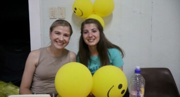 Two girls are sitting with yellow, smiley balloons in front and at the back of them. The light-hair girl is wearing brown, the dark-hair girl is wearing a greenish shirt. The table cloth has white and red squares.