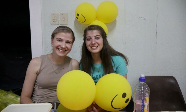 Two girls are sitting with yellow, smiley balloons in front and at the back of them. The light-hair girl is wearing brown, the dark-hair girl is wearing a greenish shirt. The table cloth has white and red squares.