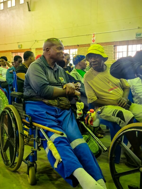 Two men seated at the event, one using a wheelchair, talking and smiling among other attendees in a hall.