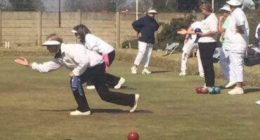 An Image of two people bowling and other people standing at the back wearing bowling clothing at the Vryheid Bowling Club.