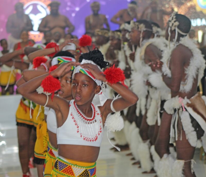 A group of young dancers, dressed in their vibrant traditional attire, performing energetically. A girl in the foreground wears a beaded top with colourful patterns, with several other dancers seemingly following her lead.