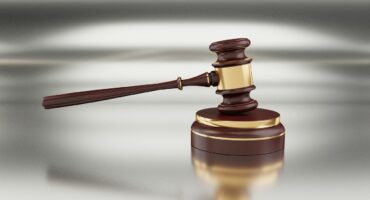 A wooden judge’s gavel with gold accents resting on a matching sound block against a blurred metallic background.