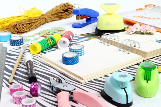Scrapbooking supplies arranged on a table, including an open blank notebook, coloured tapes, glue sticks, craft punches, string, scissors and decorative elements.