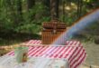 A brown picnic basket is on a table. The table has a white and red squared pattern. There is a small, greet bucket in the foreground. There is a blurry forest in the background and a rainbow across the picture.