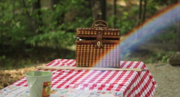 A brown picnic basket is on a table. The table has a white and red squared pattern. There is a small, greet bucket in the foreground. There is a blurry forest in the background and a rainbow across the picture.