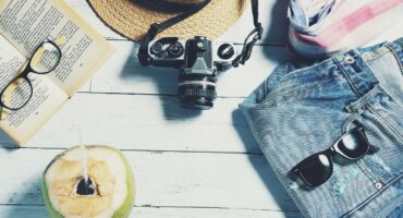 Holiday essentials laid out on a wooden surface, including a camera, hat, book with glasses, coconut drink, scarf, denim shorts and sunglasses.