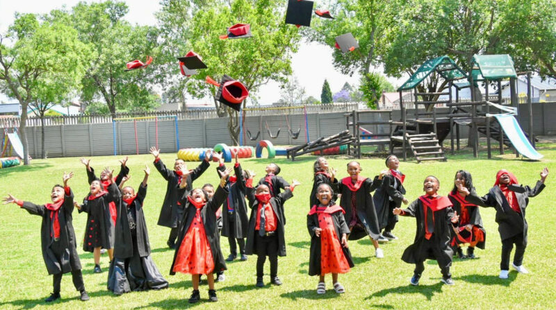 A group photo of Wonderland Pre-Primary School's Graduating class. Children are dressed in their black and red graduation gowns throwing caps in the air.