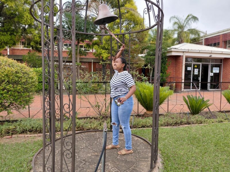A photo showing learners from the Bridging Group 2025 taking turns to ring a ceremonial bell outdoors at Tugela Pre-Vocational School. Each photo features one or a small group of learners standing beneath the metal bell structure in a garden area with trees and school buildings in the background.