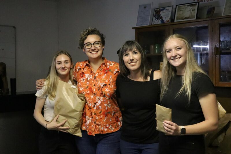 There are four people. The first woman has a brown paper bag in her hand. The second woman is wearing orange with glasees. The third and fourth women are wearing black. The background is dark.