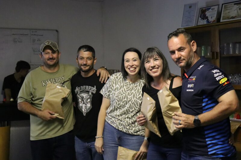 There are five people. Person one, four and five have brown paper bags in their hands. person one is wearing a khaki shirt and cap. Person two and four is wearing black. Person three is wearing green. Person five is wearing a Redbull racing shirt. There is a white wall in the background.