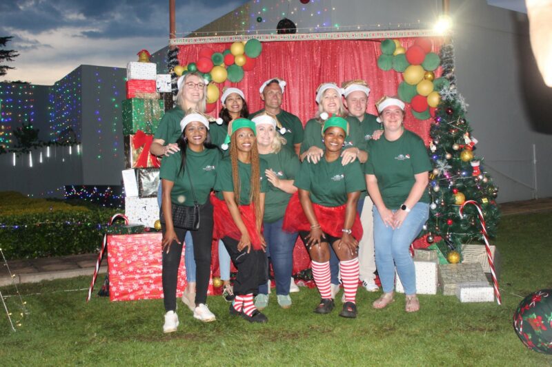 A group of people dressed in Christmas attire pose for a photo in front of a Christmas backdrop.