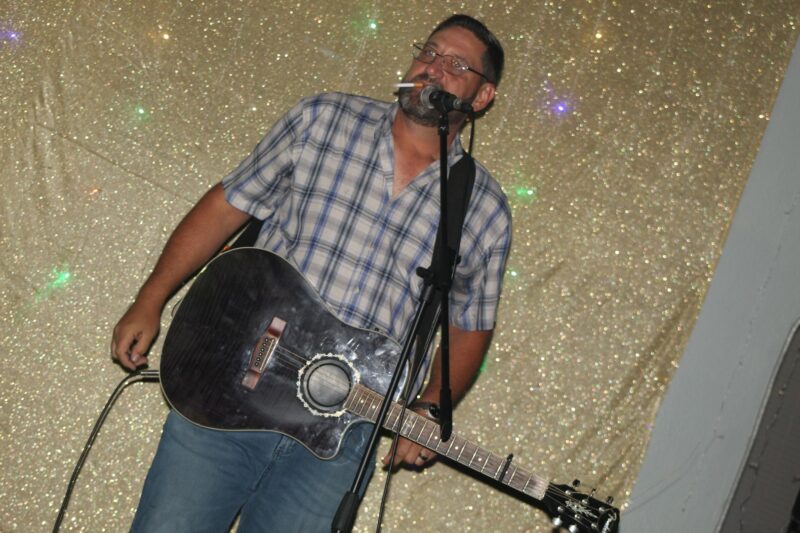 A man with a guitar performing in front of a golden glitter backdrop.