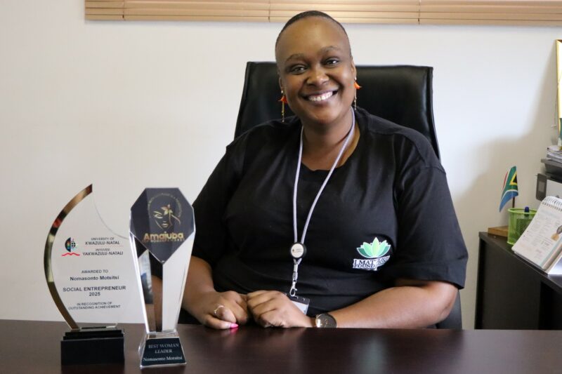 A woman is smiling. She is sitting behind a desk on a black chair. There are two trophies on the table with wording on them. She is wearing black with green printing in the front. There is a chain around her neck. The wall behind her is white.