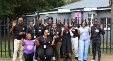 Group of smiling adults wearing foundation-branded shirts standing and waving together outside a small grey building.
