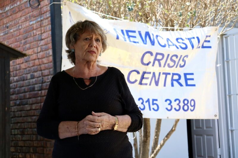An elderly lady standing in front of a white banner with blue and purple writing on it. She is wearing black. Her hands are in front of her body. It is day-time. THere is a brick wall in the background.