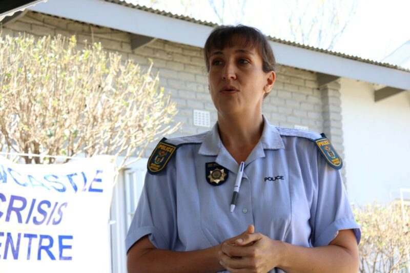 A female in a South African Police Service uniform. She is standing outside, with her hands folded in front of her chest. There is a brick wall and a white banner in the background. It is day-time.