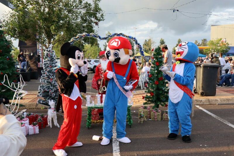 Three cartoon looking characters are standing around. It is day time. There are Christmas decorations in the background. The characters are wearing blue, white, red and black. One looks like Mickey Mouse. The other character looks like Mario from Mario Cart. The blue character looks like a cat. They are standing in the Newcastle Mall parking lot.