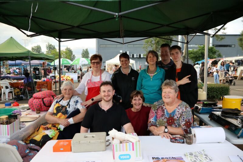 A group of people standing under a tents. They are baking pancakes. THey are wearing different colour clothing. It is day-time. There are other market stalls in the background.