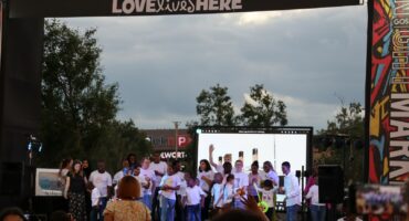 A stage with children on it. The children are clothed in whote. It is dawn. THere is a screen behind the stage. THere is a cross-bar over the stage with words "Love shines here." There are people sitting on white chairs facing the stage.