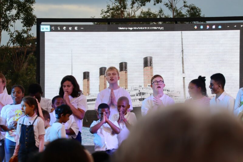Children are standing on a stage, praying with their eyes closed. There is a screen behind them with an image that looks like a ship (Titanic). The picture has a white background. There are lights shining on the children. The children are different lenghts and ethinicities.