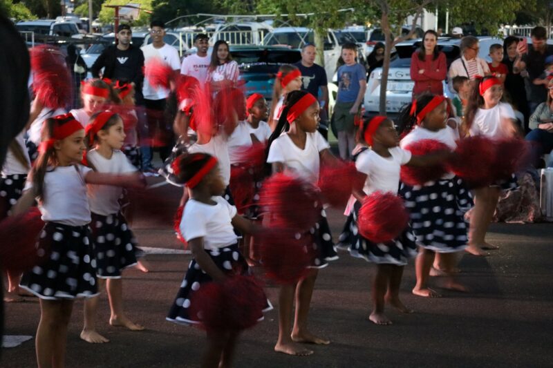 Primary school children dancing in a group. They are wearing red and white with red pom-poms in their hands. The children are wearing red ribbons in theur hair. The children are barefoot and standing on tarmac. It is night time.