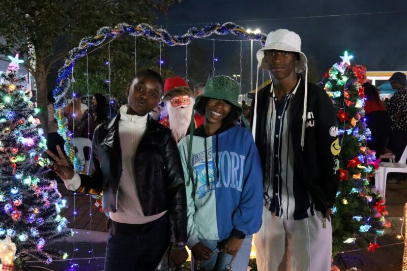 Three people standing in front of a Santa Clauss statue. THere are lights in the form of a heart. It is night time. Many lights of different colours are visible in the background. Two of the three people are wearing hats on their heads.