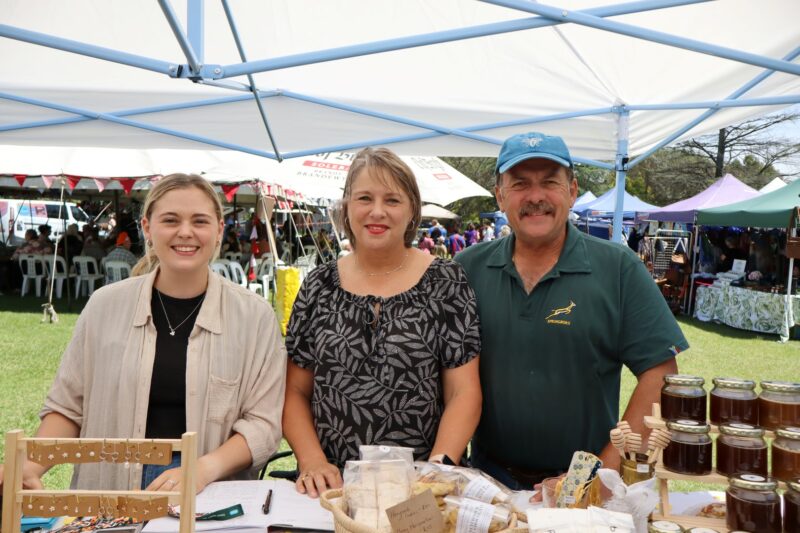 Three people are facing the camera. The man has a blue cap on his head. He is wearing a green Springbok rugby jearsey. They are standing under a shite tent. THere are various products in front of them on the table.