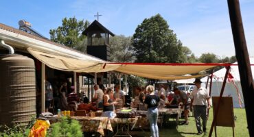 People browse stalls under shade canopies at an outdoor Christmas market beside a church building.