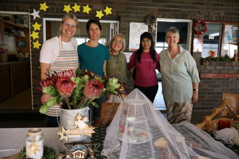 Five women pose behind a decorated Christmas stall with flowers and baked goods.