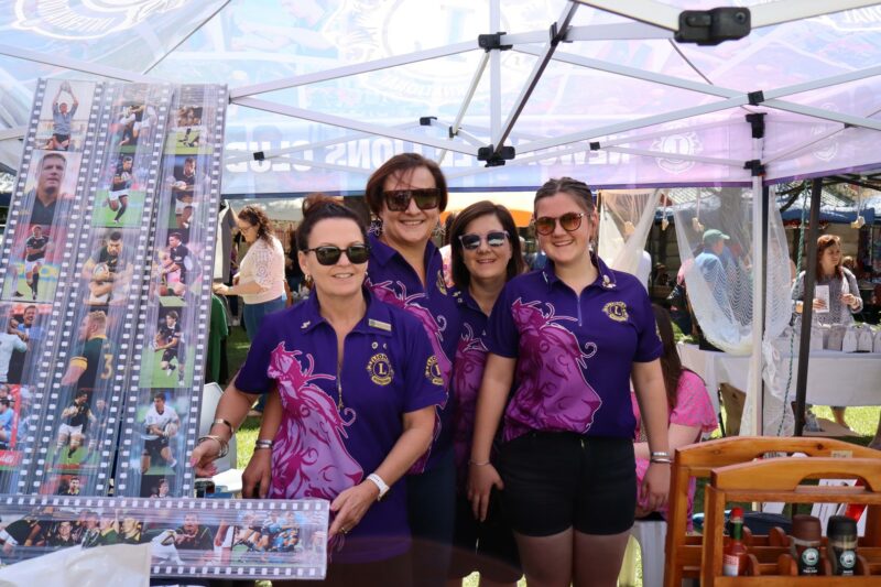 Four woman dressed in purple is standing under a tent. Al of them are wearing sunglasses. There is a big portrait in front of them. They are wearing black pants.