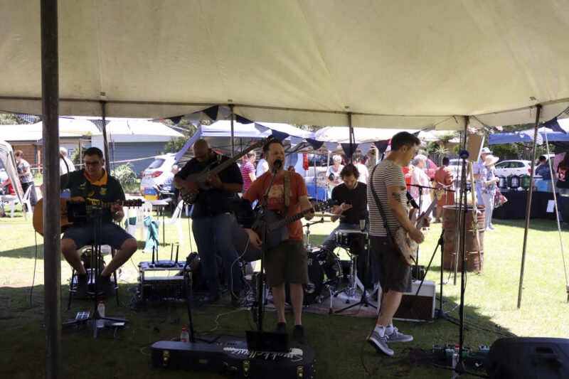 A band of 5 people playing various instruments under a big, white tent. THere are guitars and a drum set. A man is singing. there are many cables. The drum set is on a carpet. It is day-time. Besides the drummer, there is only one guitarist sitting.