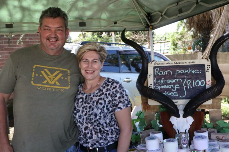 A man and a woman is smiling. There is a car behind them. THere is a board with chalk writing on it. THere is a skull of an animal with horns. The people are standing under a green tent.