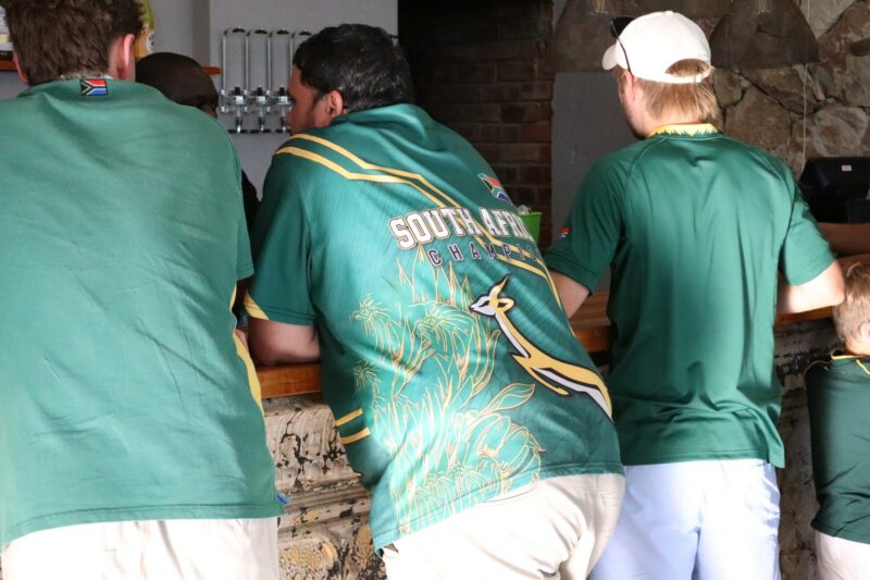 Three men standing with their backs to the camera. They are wearing green and Springbok jerseys. One man is wearing a white cap.