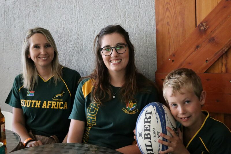 Three people are wearing Springbok rugby jerseys. A small boy is holding a rugby ball in his hands. There is a grey wall and wooden door in the background.