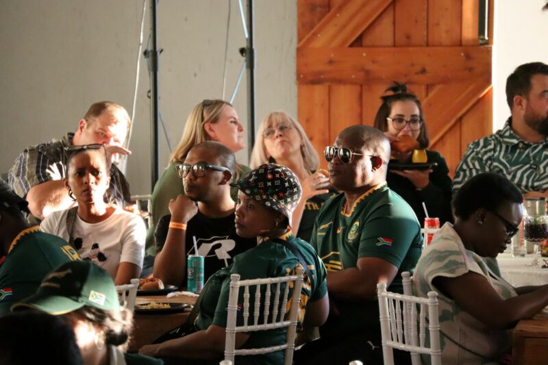 People sitting on white chairs staring in front of them. They are indoors. They are wearing Springbok rugby jerseys. A woman has a hat on her head. THere is a grey wall and wooden door in the background.
