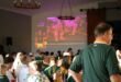 Crowd of rugby supporters in green jerseys watching a Springboks match projected on a large screen inside a hall.