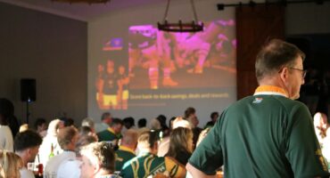 Crowd of rugby supporters in green jerseys watching a Springboks match projected on a large screen inside a hall.