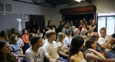 A large group of people sit closely together in a brightly lit room, watching an event or presentation, with more people standing at the back.
