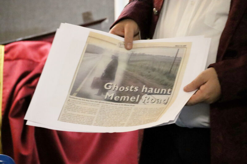 A man is holding a newspaper article in his hands. It looks like he is showing it to people. On the paper it shows a ghostly figure. There is a red robe in the background.